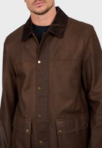 Chaqueta de cuero marrón con cuello de pana, cierre frontal a presión, dos bolsillos delanteros y textura suave. Los detalles de diseño clave incluyen costuras.
