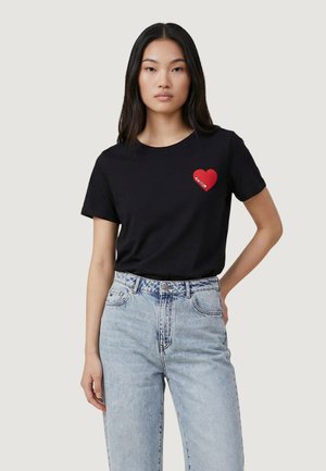 Giovane donna con lunghi capelli neri indossa una T-shirt nera con cuore rosso e scritta "amour", abbinata a jeans a vita alta azzurri.