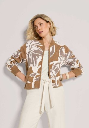 Frau mit kurzen blonden Haaren trägt eine braun-weiße, mit Blumen gemusterte Strickjacke und cremefarbene, hoch taillierte Hose mit Bindegürtel.