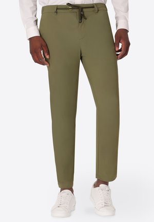 Uomo che indossa pantaloni slim verde oliva con coulisse in vita, camicia bianca e sneakers bianche, in piedi davanti a uno sfondo chiaro.