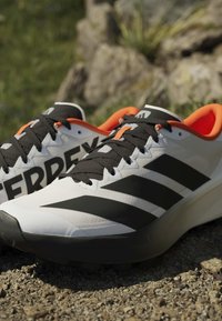 Chaussures de trail Adidas Terrex blanches et noires avec intérieur orange, posées sur un sol extérieur rocheux.