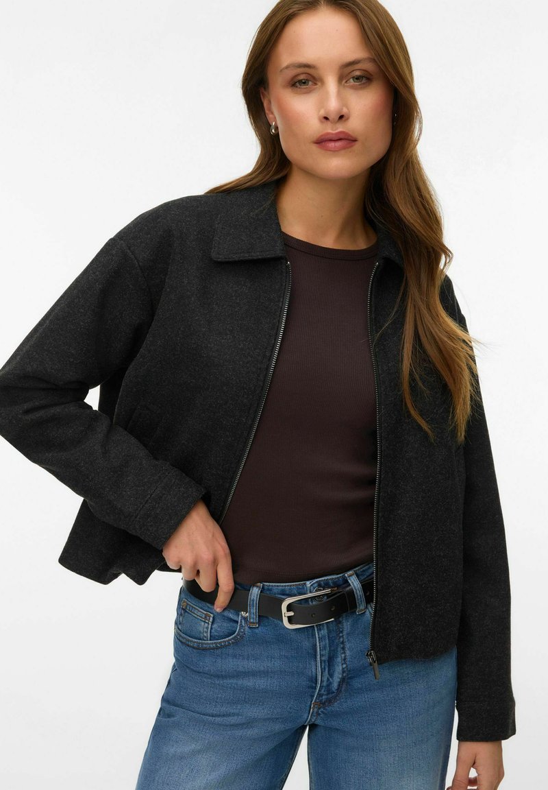 Femme aux cheveux longs portant une veste zippée gris foncé sur un haut marron et un jean bleu avec une ceinture noire, posant devant un fond blanc.