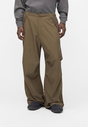 Persona che indossa pantaloni cargo marroni larghi con tasca laterale con zip e scarpe nere su sfondo semplice.