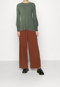 Groene gebreide trui met ribbeltextuur, gecombineerd met losse bruine wijde broek. De schoenen zijn zwarte platformschoenen met een stevige zool.