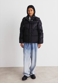 Mustat puffatakki, jossa on vetoketjukiinnitys, irrotettava huppu ja suuret tilkkupaneelit. Yhdistetty vaaleansinisillä, haalistuneilla leveälahkeisilla farkuilla ja mustilla kengillä.