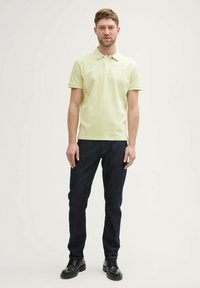 Polo shirt jaune clair avec col et manches courtes, associé à un jean bleu foncé et des chaussures noires. Design simple et ajusté.