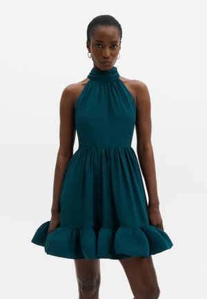 Vrouw die een mouwloze teal halterjurk draagt met een geplooide hoge kraag en een volumineuze geplooide zoom, staand tegen een effen achtergrond.