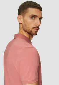 Kurzarm-Polohemd in staubrosafarbe, aus Baumwolle, mit einem Kragen und subtiler Textur. Modell zeigt das Profil von der Seite.