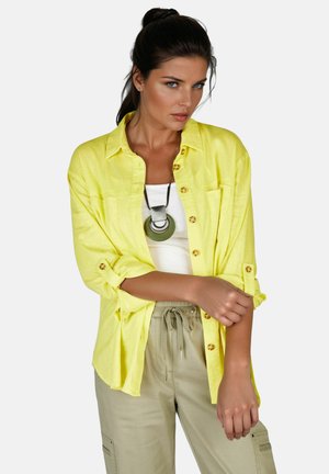 Kvinde med mørkt hår iført gul skjorte med knapper over hvid top, beige bukser med bindebånd og stort rundt vedhængs halskæde, poserer.