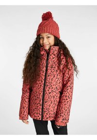 Veste en duvet rose avec un motif à taches noires, col montant et fermeture éclair ; portée avec un bonnet en tricot rouge avec un pompon sur le dessus.