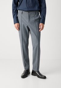 Muž v šedých kostkovaných kalhotách, černých kožených botách a tmavě modré košili s dlouhým rukávem stojí na bílé podlaze.