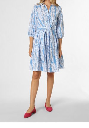 Mujer con vestido azul claro con estampado de hojas blancas, con botones al frente, cintura atada, mangas tres cuartos y zapatos rojos de tacón bajo tipo bloque.