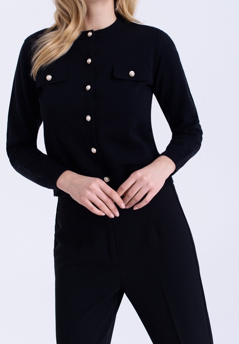 Femme portant un cardigan noir boutonné avec des boutons en perle et un pantalon noir assorti, posant les mains croisées à la taille.