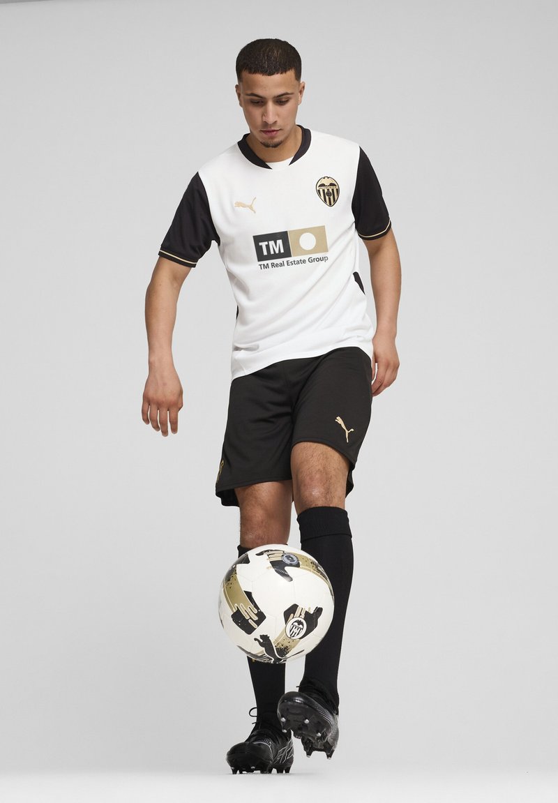 Valge ja must jalgpallipaita kuldsete aktsentidega, mustad lühikesed püksid ja mustad sokid. Mängija seisab jalgpalliga jalgade vahel halli tausta taustal.