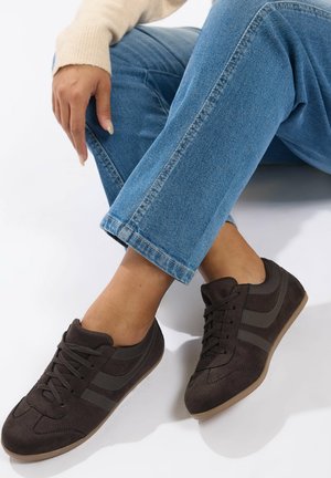 Nainen, jolla on mustat mokkatennarit ja siniset farkut, istuu valkoista taustaa vasten, toinen käsi polvella lepäämässä.