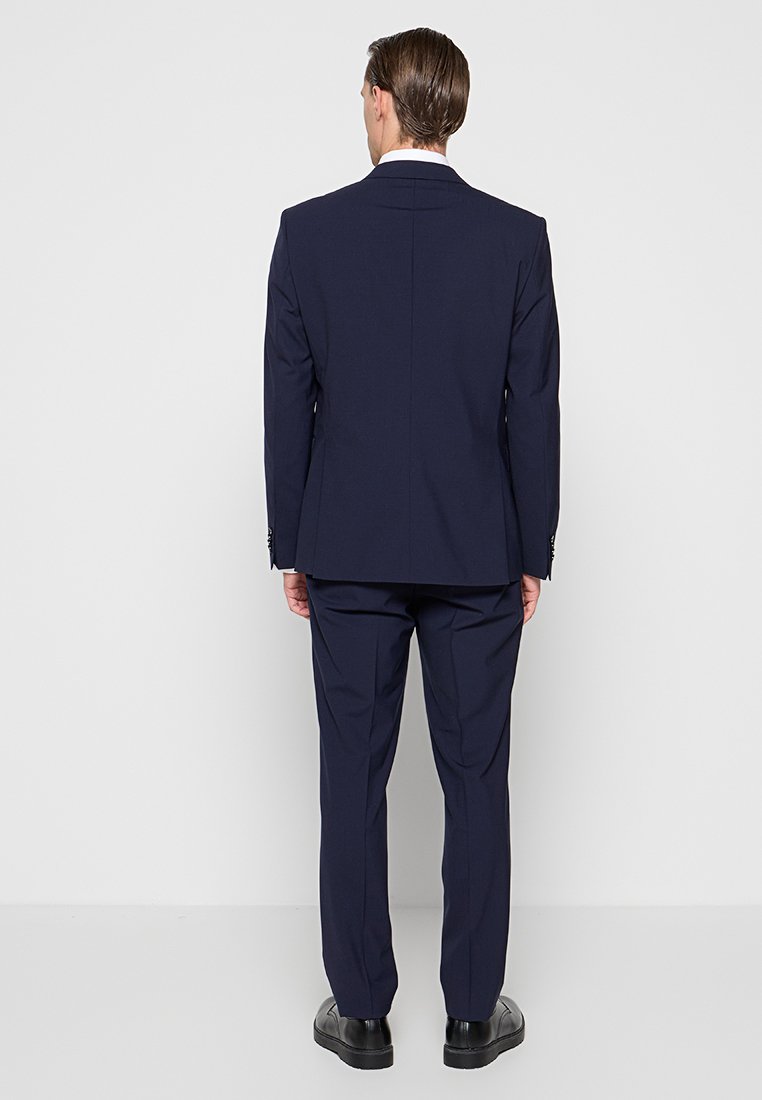 Mann steht mit dem Rücken zur Kamera, trägt einen marineblauen Anzug, der weiße Kragen des Hemdes ist sichtbar, und schwarze Anzugschuhe vor einem schlichten grauen Hintergrund.