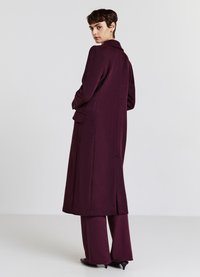 Donna con capelli corti che indossa un lungo cappotto viola scuro e pantaloni larghi abbinati, mentre guarda oltre la spalla su uno sfondo neutro.