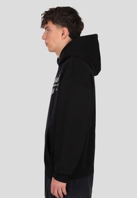 Hanorac negru cu glugă și croi lejer, având un buzunar frontal și mansete reiate. Textură moale, design simplu fără modele vizibile.