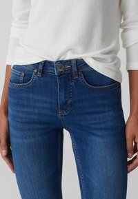 Jeans in denim blu con vita media, design a cinque tasche e chiusura con bottone metallico. Il tessuto ha una texture liscia con una leggera scoloritura.