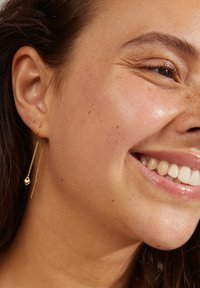 Boucles d'oreilles en or avec une chaîne fine présentant une perle ronde et une barre droite, pendent élégamment près de l'oreille du modèle. Texture lisse.