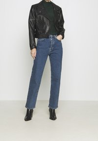 Chaqueta biker corta de cuero negro sobre una blusa verde con textura, combinada con vaqueros azules de corte recto y botines negros de tacón.