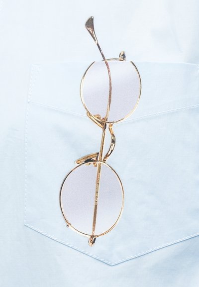 Στρογγυλά γυαλιά με χρυσή περίμετρο κρεμασμένα από την τσέπη ενός ανοιχτόχρωμου μπλε πουκάμισου με εμφανείς ραφές.