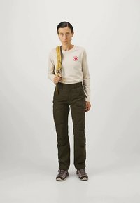 Világos bézs hosszúujjú póló logóval, sötétzöld cargo nadrág és szürke túracipő. A személy egy sárga hátizsák pántját tartja.