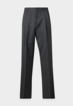 Homme portant un pull à col roulé marron foncé, un pantalon gris foncé et des mocassins marron et beige, debout avec une main dans la poche.