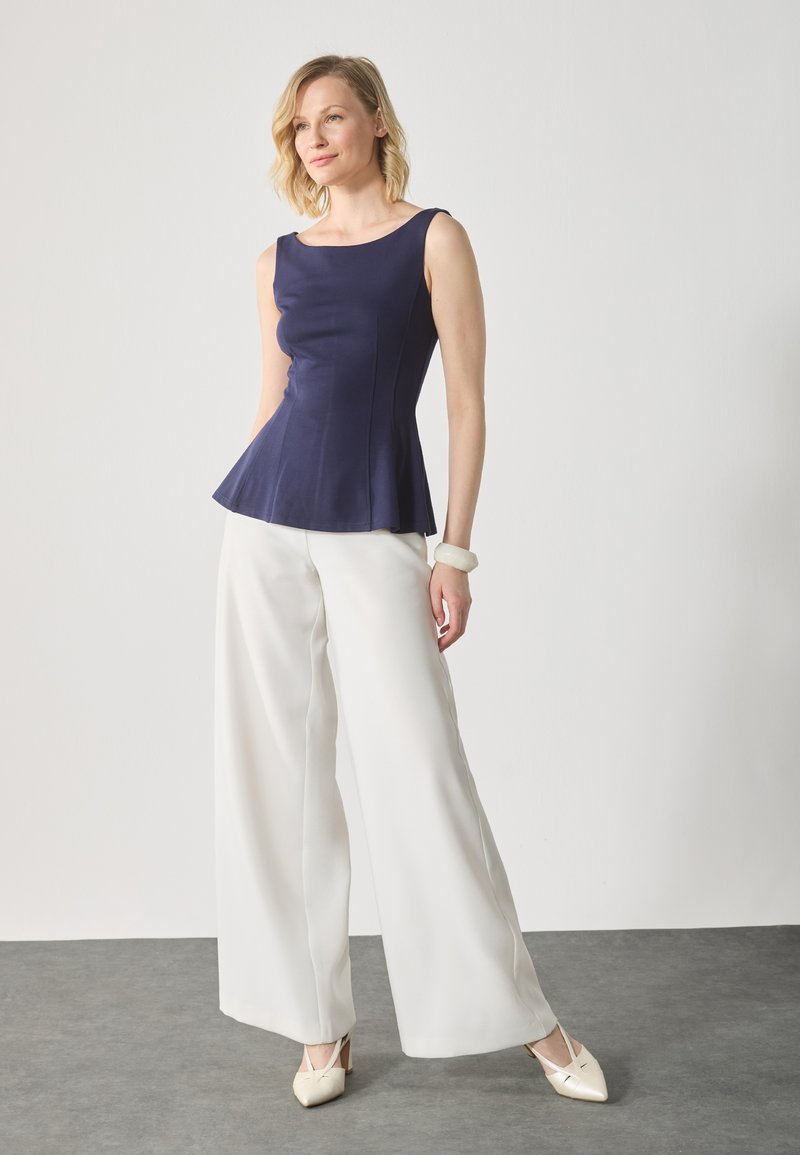 Top peplum blu abbinato a pantaloni bianchi a gamba larga. Il top presenta una texture liscia e un design leggermente svasato, completato da tacchi color crema.