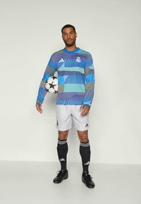Adidas langermet trøye i blå og grå geometriske mønstre, kombinert med hvite shorts, svarte sokker og fotballsko, holder en ball.
