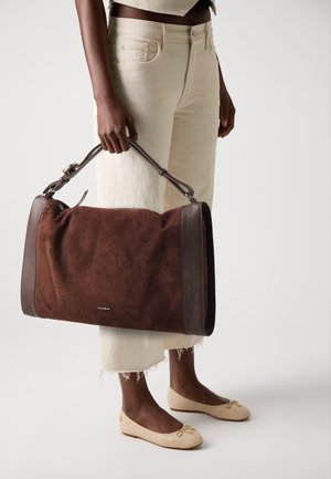 Brown suede handbag with leather accents, rectangular shape, adjustable strap, and logo detail. Model wears light-colored cropped pants and flats.