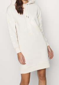 Krämfärgad hoodieklänning med en stor ton-i-ton-logga, ribbade ärmslut och en känguruficka, tillverkad av ett mjukt, texturerat tyg.