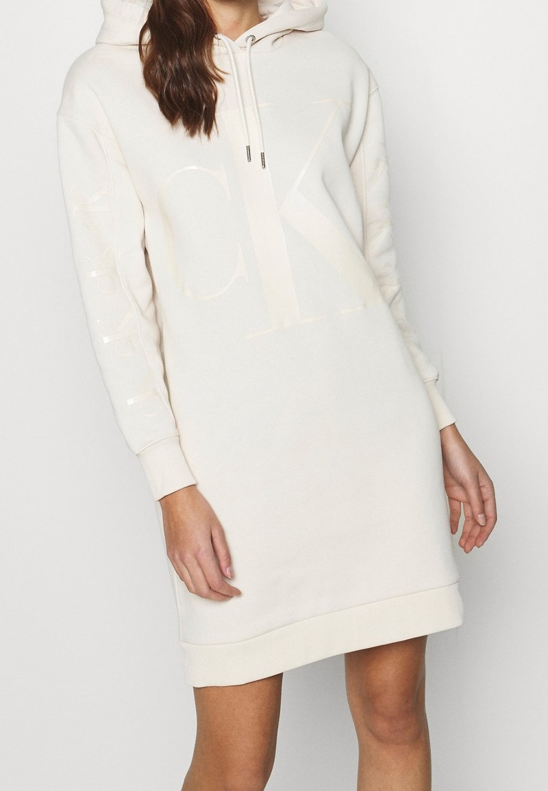 Krämfärgad hoodieklänning med en stor ton-i-ton-logga, ribbade ärmslut och en känguruficka, tillverkad av ett mjukt, texturerat tyg.