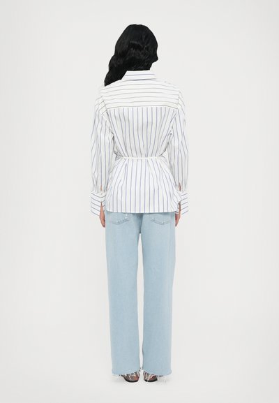 Camicia a righe bianche e blu con vita attillata, abbinata a jeans blu chiaro a gamba larga. La camicia ha polsini con bottoni e uno sprone sulla schiena.
