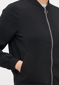 Black bomber jacket with a zip front, ribbed cuffs, and side pockets. Smooth fabric with a fitted design. Zipper features a circular pull tab.
