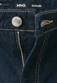 Jean en denim bleu foncé avec une fermeture éclair argentée partiellement ouverte, un bouton métallique et une étiquette de marque MNG visible à l'intérieur de la ceinture.