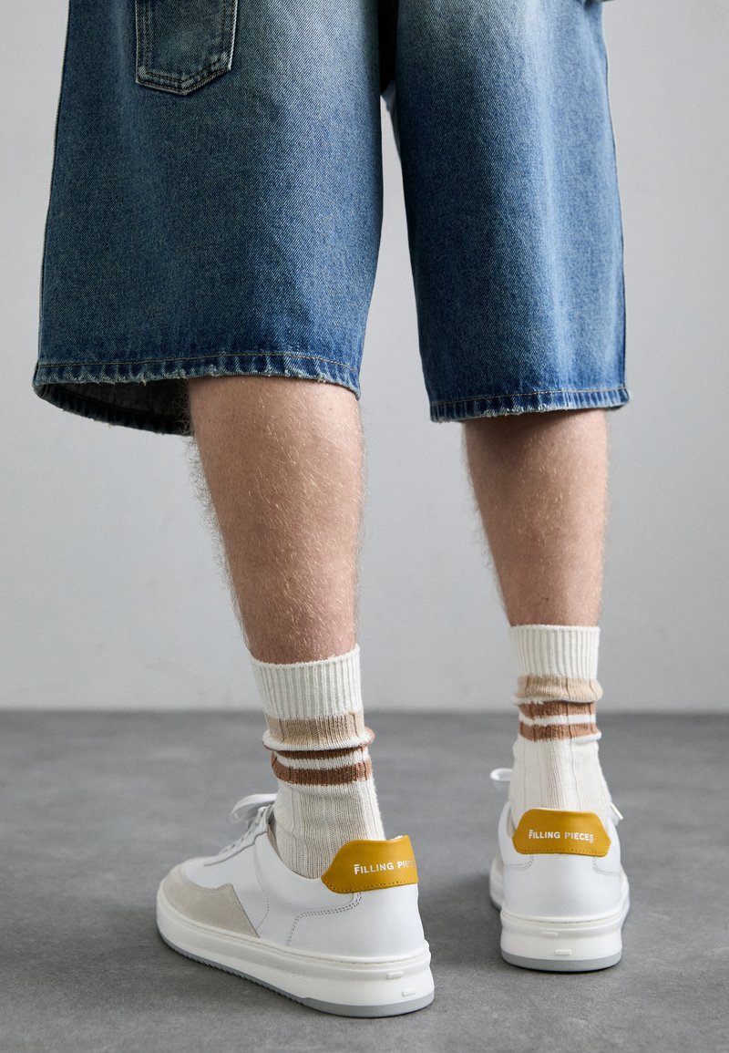 Hvite sneakers med gule detaljer, brukt sammen med kremfargede og brune stripete sokker, kombinert med blå denimshorts. Glatt tekstur, avslappet design.