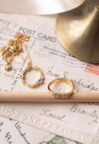 Een gouden bloemenring met pastelaccenten ligt op een houten potlood. Een bijpassende gouden bloemenketting ligt ernaast, tegen vintage ansichtkaarten.