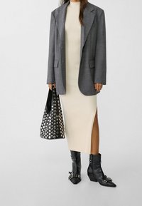 Frau trägt ein cremefarbenes, geripptes Midikleid, grauen Blazer, schwarze genietete Stiefel und hält eine schwarze genietete Handtasche vor einem schlichten Hintergrund.