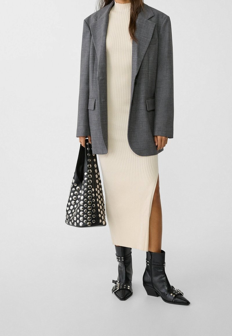 Frau trägt ein cremefarbenes, geripptes Midikleid, grauen Blazer, schwarze genietete Stiefel und hält eine schwarze genietete Handtasche vor einem schlichten Hintergrund.