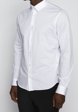 Uomo che indossa una camicia bianca aderente a maniche lunghe con bottoni, colletto appuntito e pantaloni neri, in piedi davanti a uno sfondo semplice.