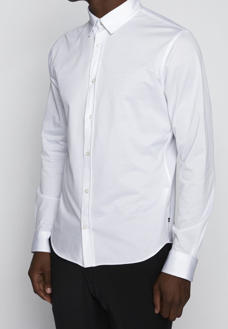 Homme portant une chemise blanche cintrée à manches longues boutonnée avec un col pointu et un pantalon noir, debout devant un fond uni.