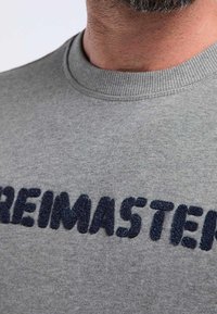 Suéter gris con cuello redondo, que presenta un grueso texto trenzado de color azul marino bordado "REIMASTERED" en el pecho.