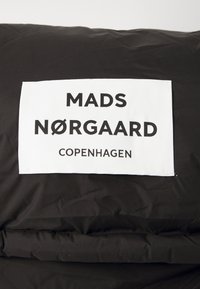 Crna puf jakna s bijelom etiketom na kojoj piše "MADS NØRGAARD COPENHAGEN" podebljanim, crnim tekstom. Glatka, sjajna tkanina s kvadratnim uzorkom.