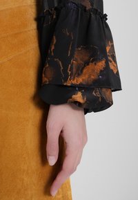 Blouse noire à motif abstrait orange, manchettes à volants, associée à une jupe en daim jaune moutarde. Montre une main reposant sur le tissu.
