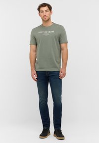 Olīvu zaļš īsām piedurknēm T-krekls ar grafisku apdruku, apvienots ar tumši ziliem pieguļošajiem džinsiem un melniem sporta apaviem. Vienkāršs un tīrs dizains.