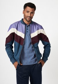Color-blocked bomber jacket in teal with purple, burgundy, and cream panels; paired with a blue geometric pattern polo shirt.