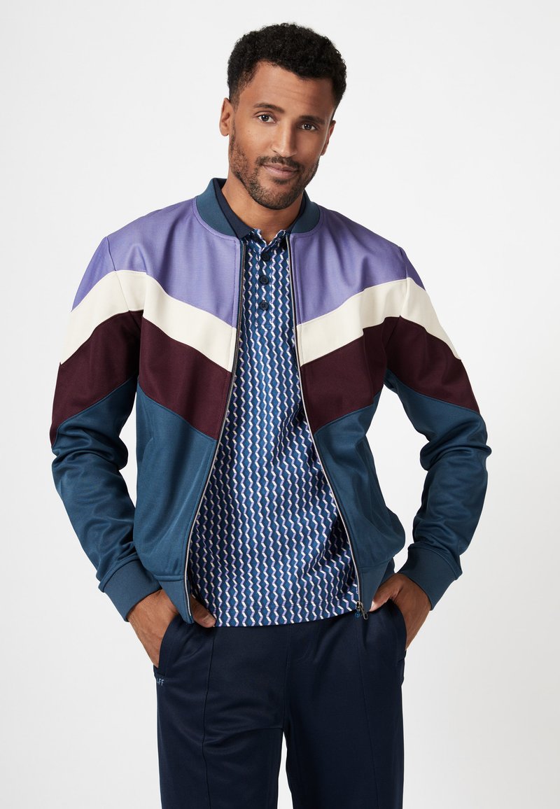 Color-blocked bomber jacket in teal with purple, burgundy, and cream panels; paired with a blue geometric pattern polo shirt.
