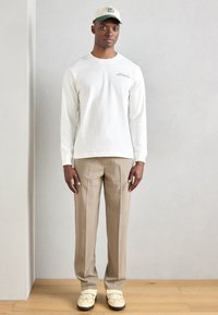 Bílé tričko s dlouhým rukávem a malým logem, béžové kalhoty a světle zelená čepice v kombinaci se sneakers s žlutými akcenty. Jednoduchý, uvolněný outfit.