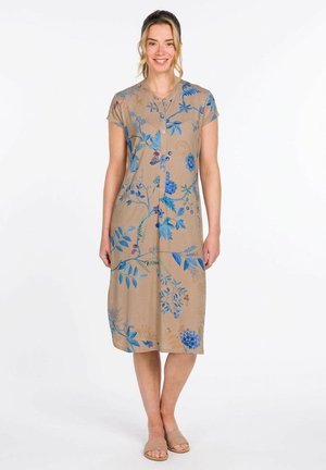 Vrouw staand, gekleed in een beige midi-jurk met korte mouwen, knopen en blauwe bloemmotieven, en beige instapsandalen.